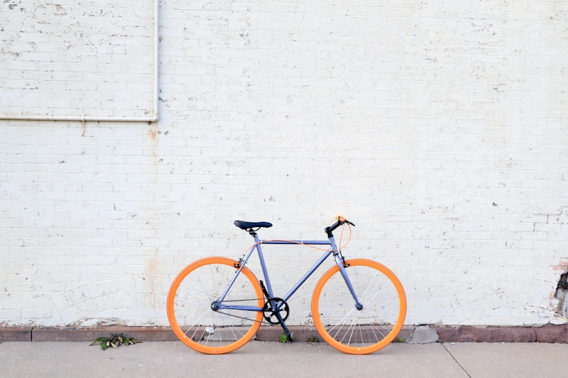 Blue-grey bicycle with orange wheels parked on a sidewalk against a white brick wall